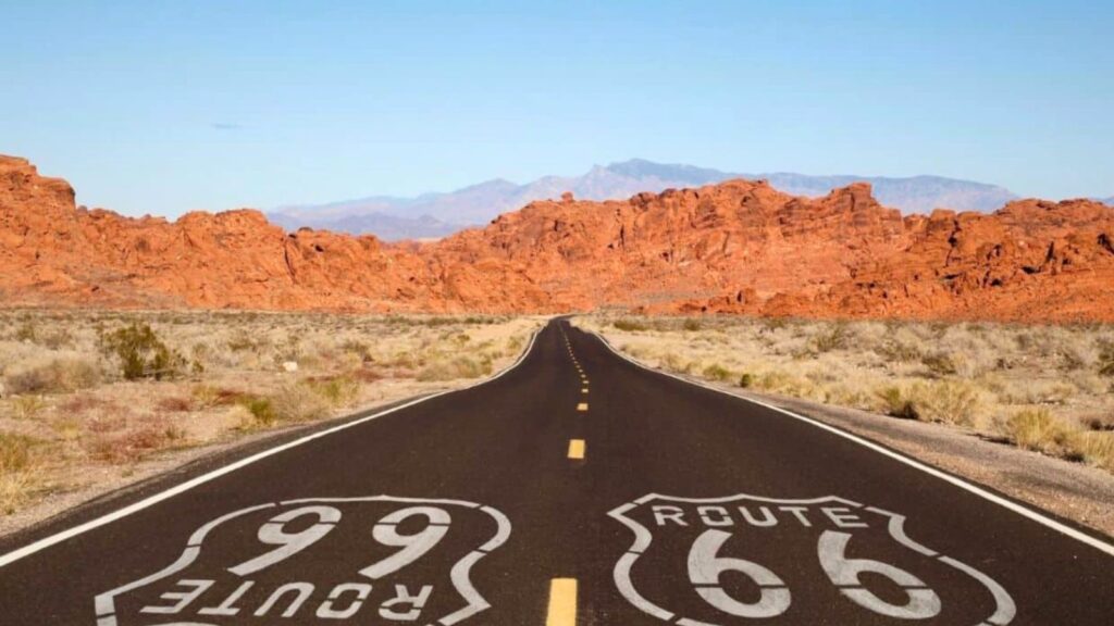 A straight highway marked with Route 66 and Route 99 cuts through desert terrain with red rock formations and distant mountains under a clear sky.