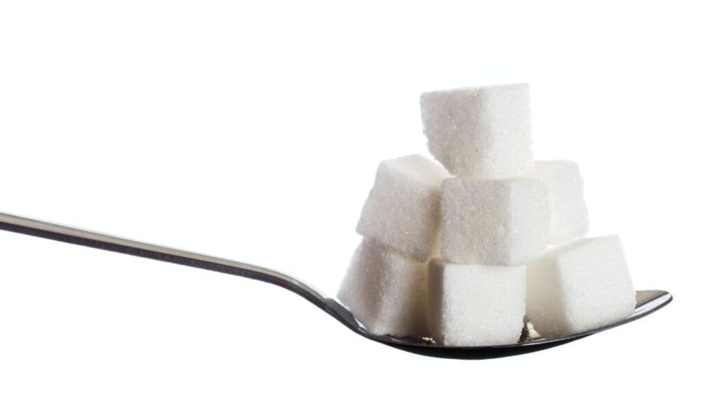 A metal spoon holds a small stack of white sugar cubes, arranged in a pyramid shape, against a plain white background.
