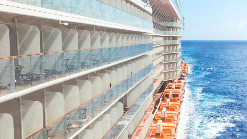 Side view of a cruise ship with rows of balcony cabins and orange lifeboats above the blue ocean, leaving a white wake behind.
