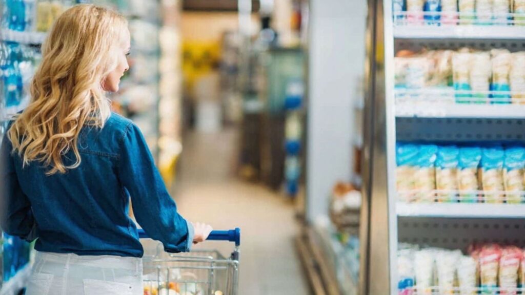 A person with long blonde hair pushes a shopping cart down a grocery store aisle, looking at shelves filled with packaged goods.