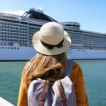 A person wearing a hat and backpack stands on a dock facing a large white cruise ship on a sunny day.
