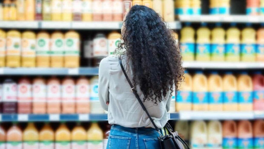 Person with long curly hair, wearing a light shirt and jeans, stands in a grocery store aisle looking at shelves filled with bottles of juice or beverages.