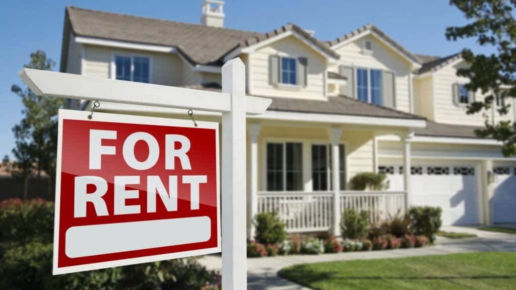 A &ldquo;For Rent&rdquo; sign is posted in front of a light-colored, two-story suburban house with a porch and a well-kept lawn.