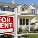 A “For Rent” sign is posted in front of a light-colored, two-story suburban house with a porch and a well-kept lawn.