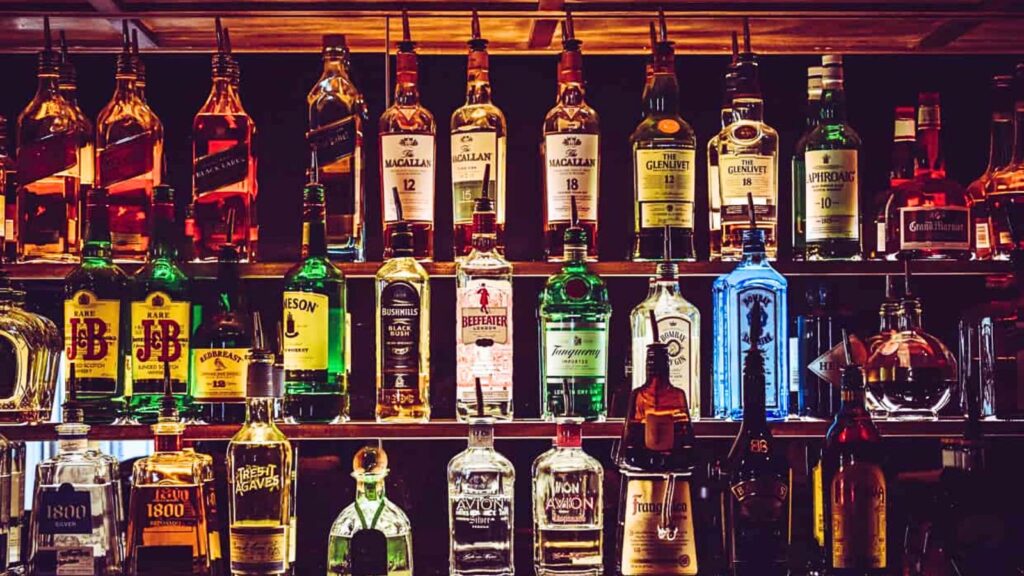 Shelves filled with various bottles of liquor including whiskey, vodka, gin, rum, and tequila, arranged in rows at a bar with dim lighting.