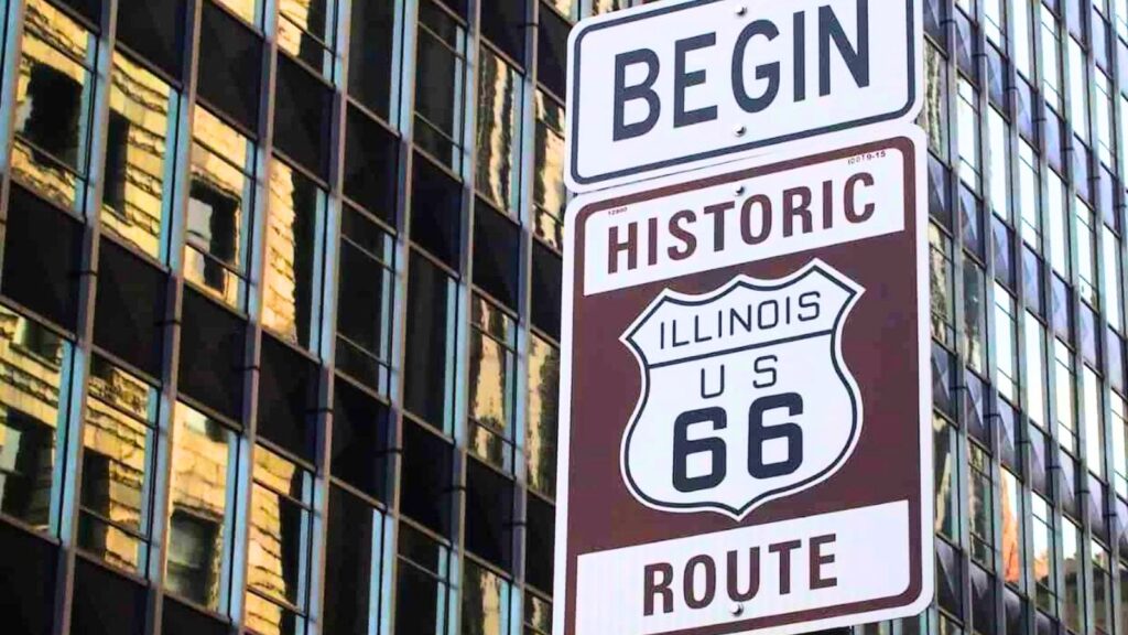 Street sign reading &ldquo;BEGIN HISTORIC ILLINOIS US 66 ROUTE&rdquo; in front of tall glass buildings, marking the start of Route 66 and celebrating Route 66&prime;s 100th anniversary.