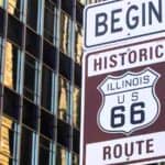 Street sign reading “BEGIN HISTORIC ILLINOIS US 66 ROUTE” in front of tall glass buildings, marking the start of Route 66 and celebrating Route 66′s 100th anniversary.