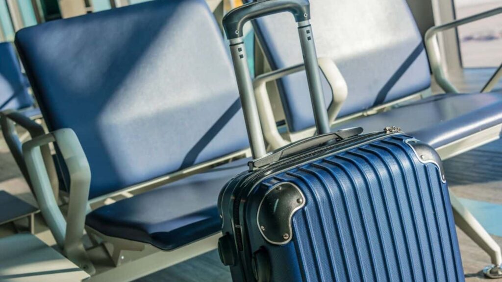 Blue hard-shell suitcase with an extended handle positioned in front of empty blue chairs in an airport waiting area, capturing a quiet moment before enjoying exclusive airport lounge perks.