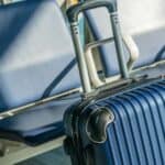 Blue hard-shell suitcase with an extended handle positioned in front of empty blue chairs in an airport waiting area, capturing a quiet moment before enjoying exclusive airport lounge perks.