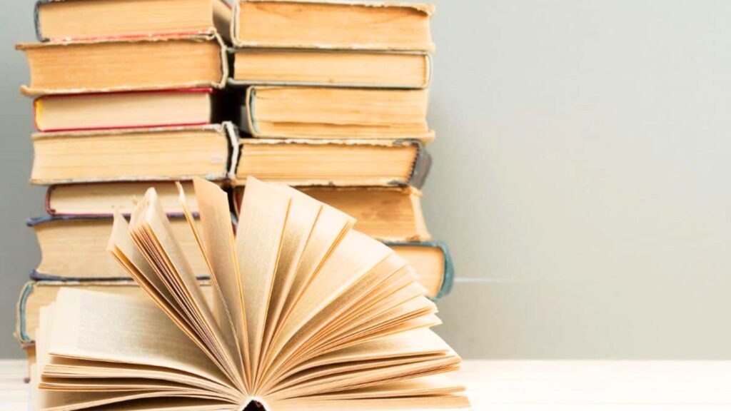 An open book lies in the foreground with stacks of closed books in the background against a plain light gray wall.