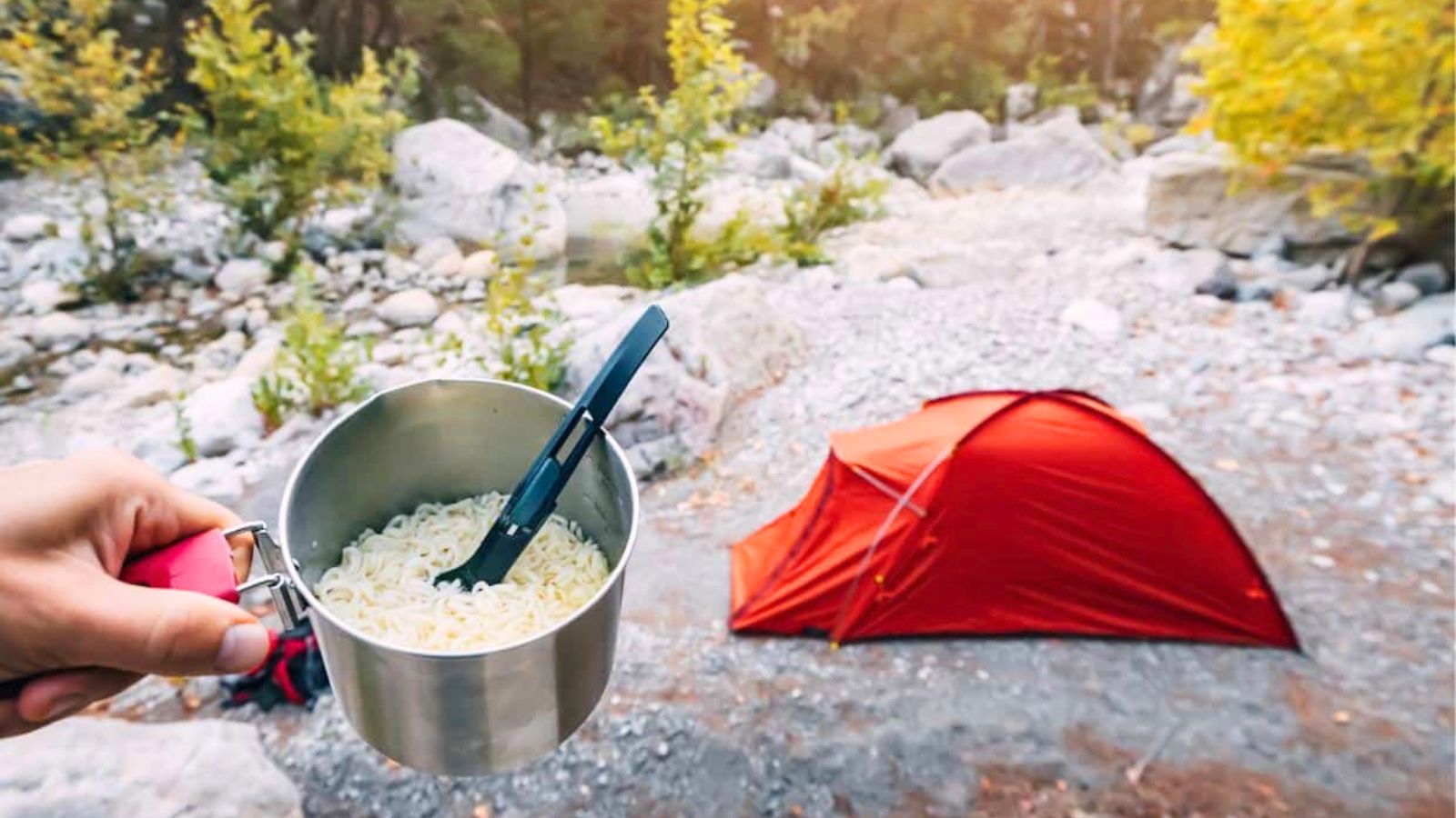 A hand holds a pot of noodles with a fork near a small red tent set up on a rocky ground outdoors with trees in the background.