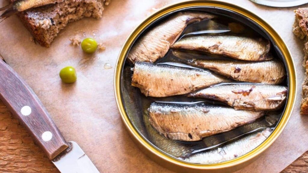 Open tin of sardines in oil with a knife, two green olives, and pieces of brown bread on a wooden surface.