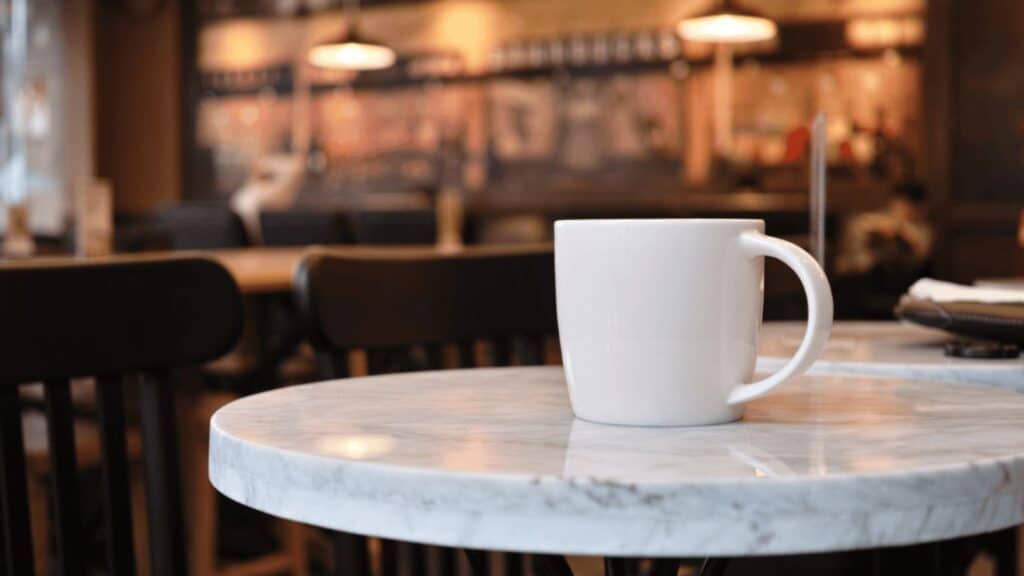 A white coffee mug sits on a round marble table in a caf&eacute;, following Italian coffee bar rules, with empty chairs and a blurred background of warm lights and wooden decor.