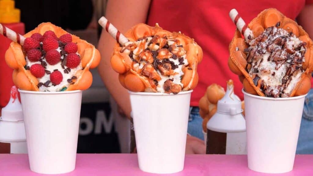 Three cups with bubble waffles filled with ice cream, each topped with different toppings including raspberries, chocolate pieces, and wafer sticks—these viral county fair foods are a feast for the senses. A person in a red shirt stands behind them.
