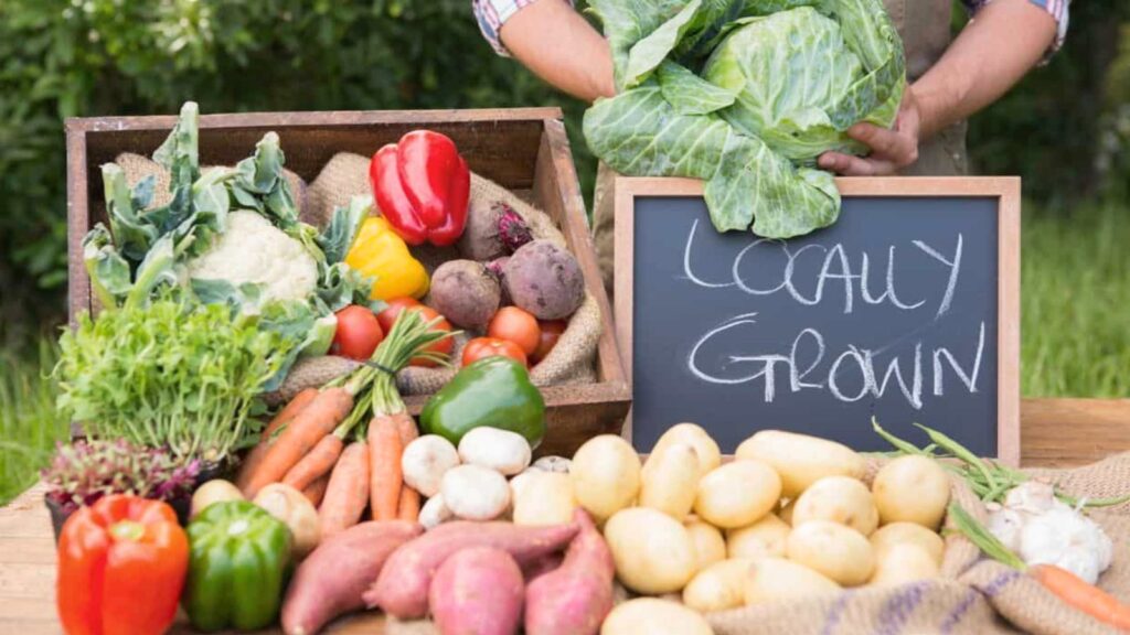 A display of fresh vegetables including carrots, potatoes, peppers, and leafy greens, with a chalkboard sign reading “Locally Grown.”.