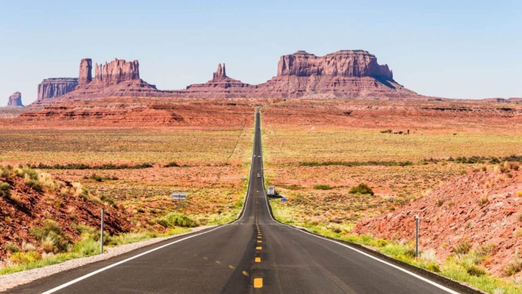 A straight, empty road stretches through a red desert landscape toward large rock formations under a clear blue sky.