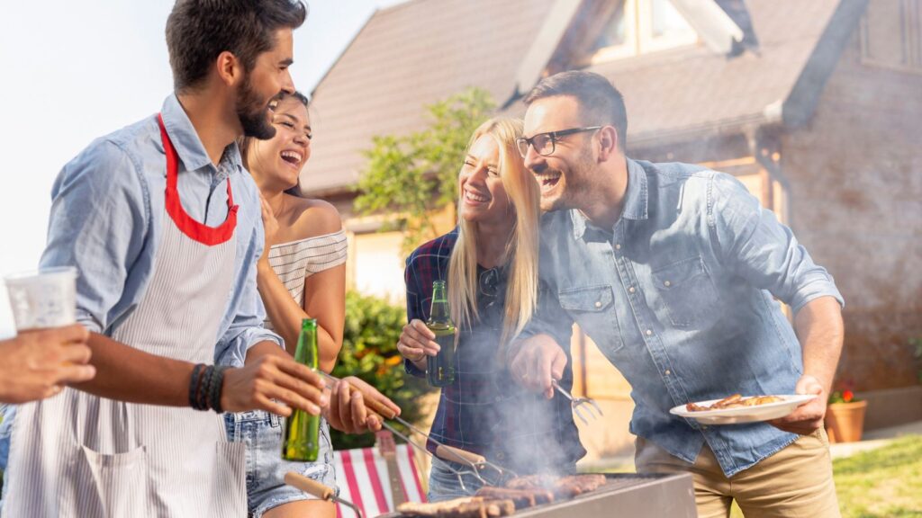Four adults stand outdoors by a grill, smiling and holding drinks, while one person cooks food. A house is visible in the background.