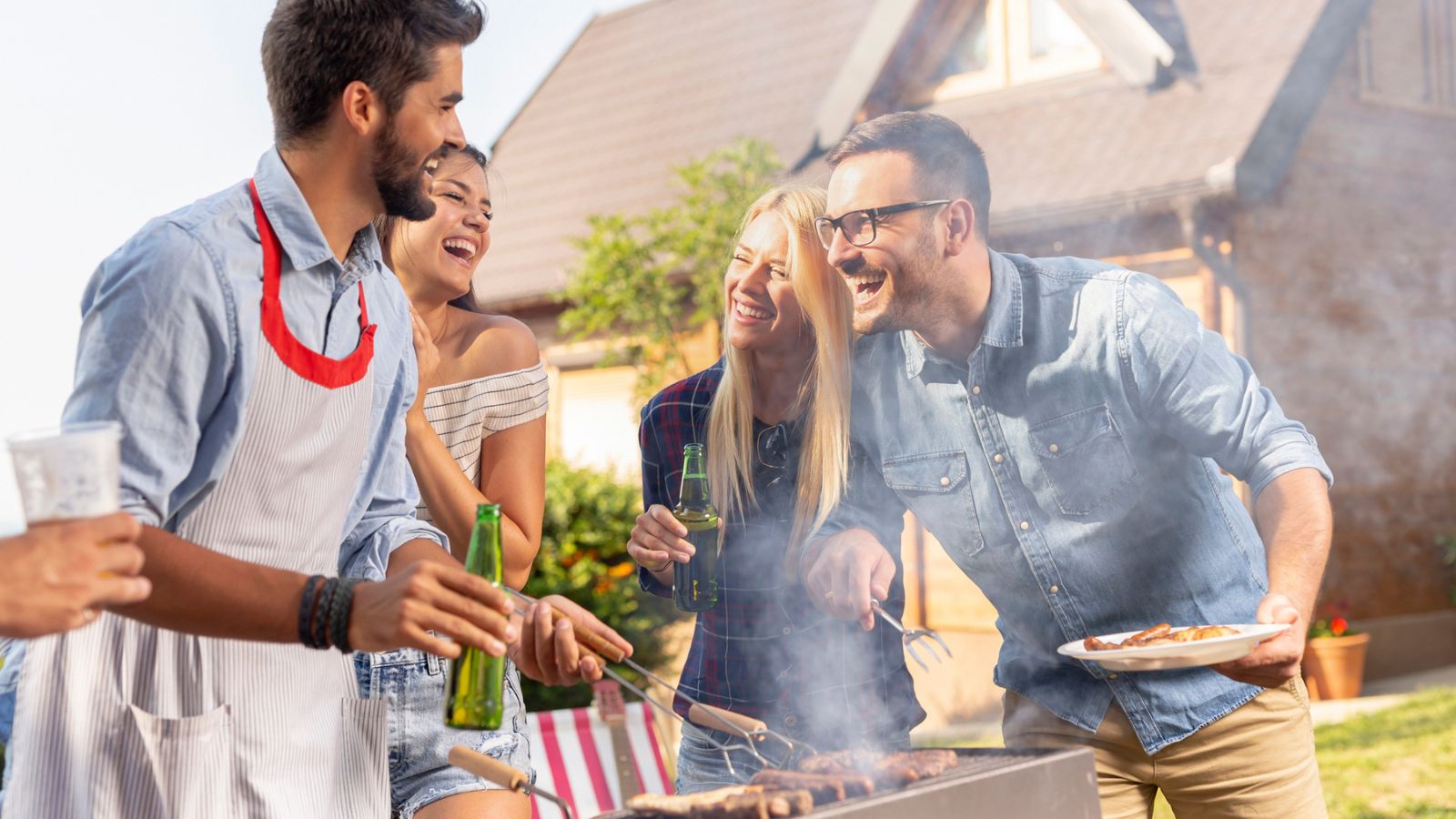 Four adults stand outdoors by a grill, smiling and holding drinks, while one person cooks food. A house is visible in the background.