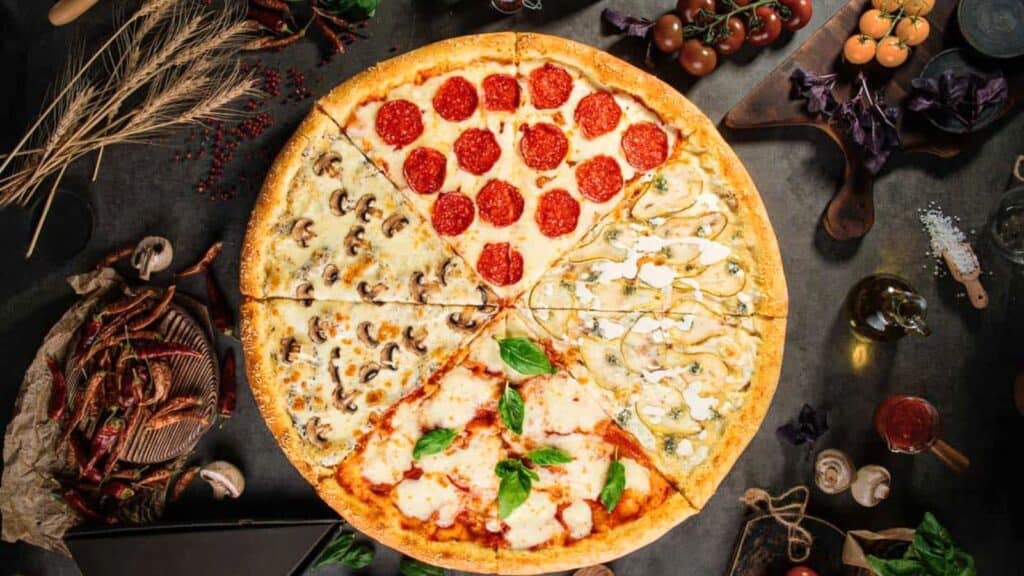 A large pizza with four different topping sections&mdash;pepperoni, mushrooms, margherita, and white sauce&mdash;is surrounded by ingredients on a dark surface.