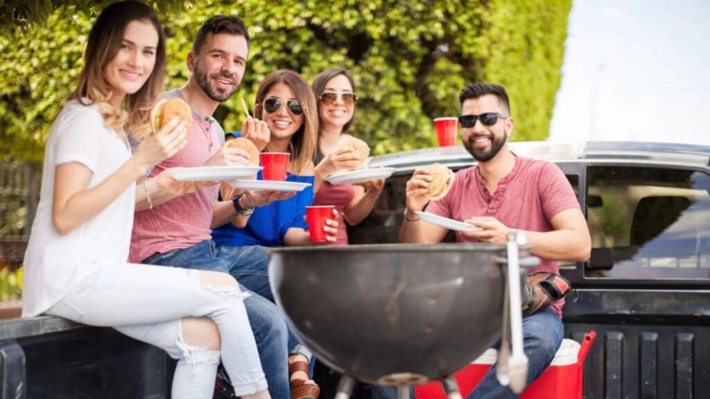 Five people sit in the back of a pickup truck holding food and drinks, smiling at the camera. A barbecue grill is in the foreground. Trees and coolers are visible in the background.