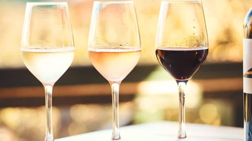 Three wine glasses filled with white, ros&eacute;, and red wine are placed side by side on a table, with a bottle partially visible on the right and a blurred background.
