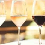 Three wine glasses filled with white, rosé, and red wine are placed side by side on a table, with a bottle partially visible on the right and a blurred background.