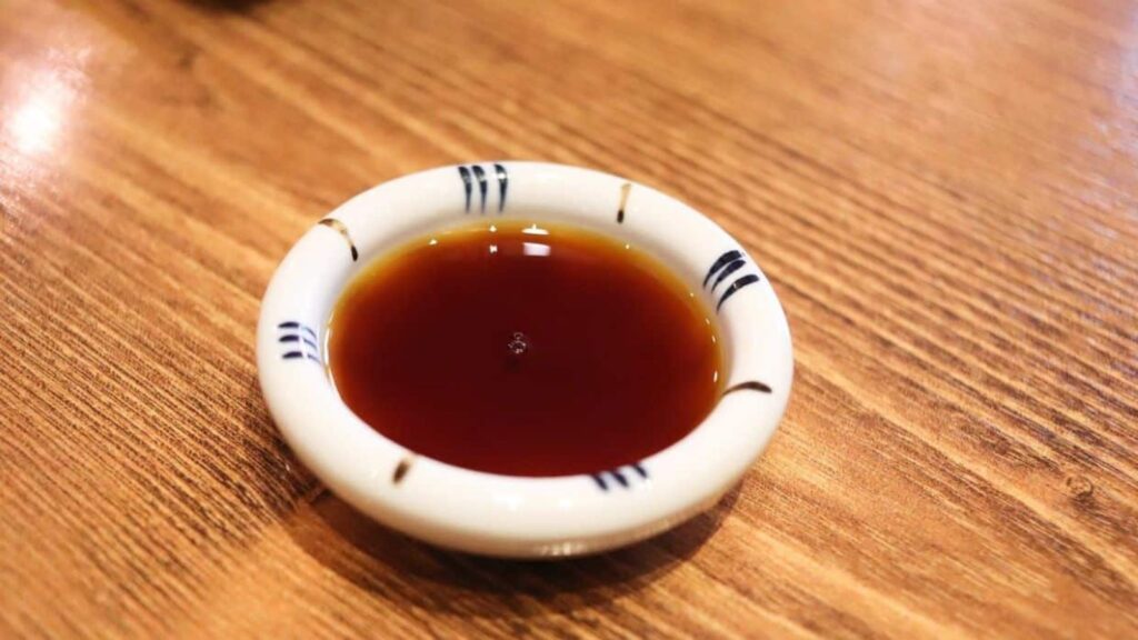 A small white dish with dark soy sauce sits on a wooden table.