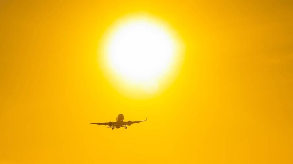 A commercial airplane flies toward a large, bright sun in a yellow-orange sky, capturing the excitement of travel during the cheapest weeks to book flights.