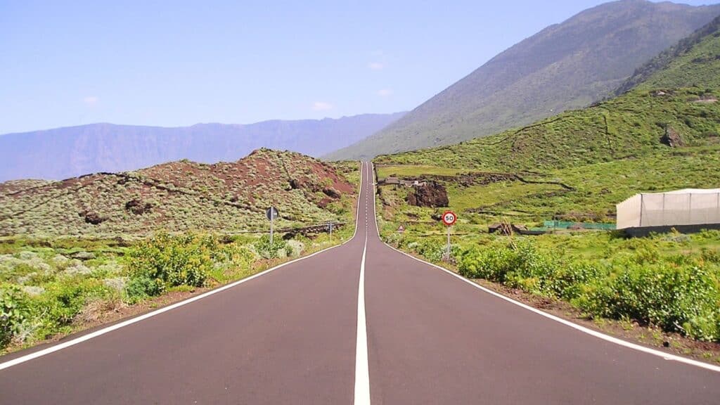 A straight road stretches into hilly, green terrain under a clear blue sky, with mountains in the background and road signs visible along the roadside.