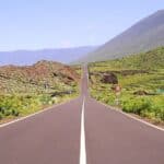 A straight road stretches into hilly, green terrain under a clear blue sky, with mountains in the background and road signs visible along the roadside.