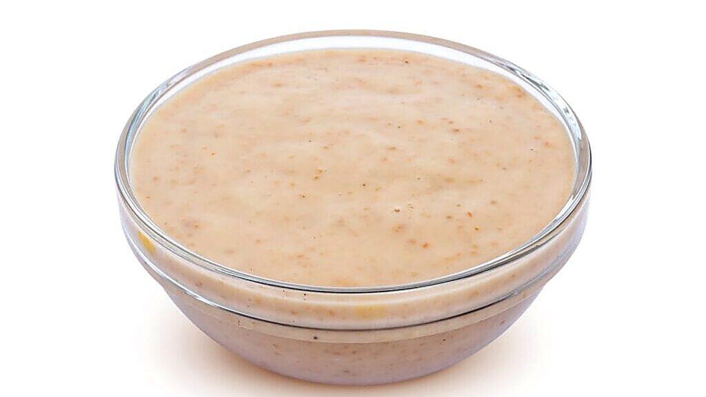 A small clear glass bowl filled with light brown creamy sauce or dressing, set against a plain white background.