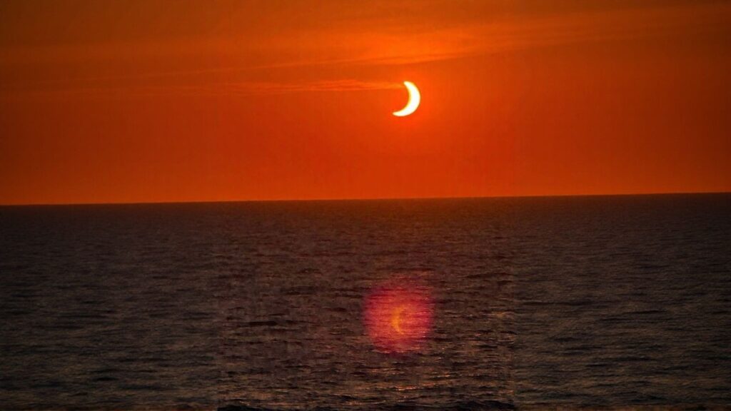 An orange sky over a calm ocean during a partial solar eclipse, with the sun appearing as a crescent and its reflection visible on the water—an unforgettable view often sought by travelers on a solar eclipse cruise.