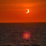 An orange sky over a calm ocean during a partial solar eclipse, with the sun appearing as a crescent and its reflection visible on the water—an unforgettable view often sought by travelers on a solar eclipse cruise.
