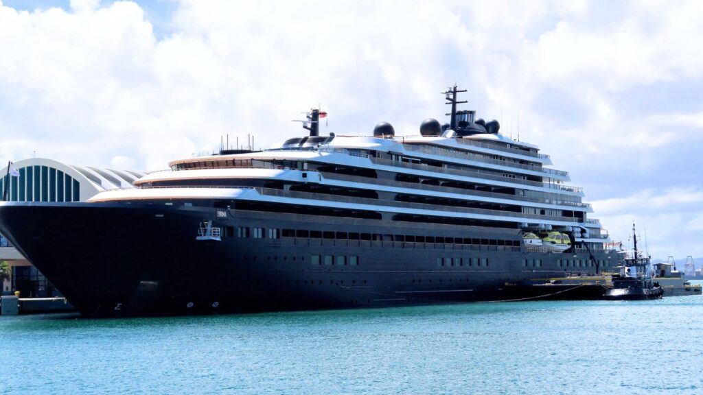 A large modern cruise ship is docked at a harbor next to a smaller vessel, with a terminal building and cloudy sky in the background.