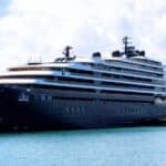 A large modern cruise ship is docked at a harbor next to a smaller vessel, with a terminal building and cloudy sky in the background.