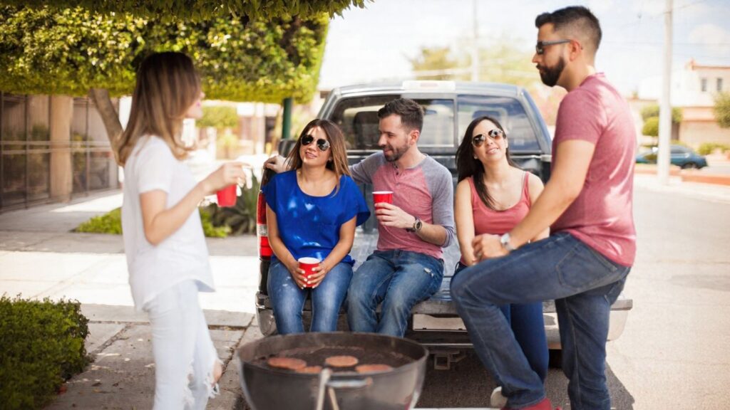 Five adults socialize by a pickup truck, holding red cups, with a grill cooking burgers in the foreground on a sunny day.