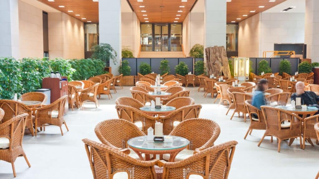 Spacious indoor cafe with wicker chairs and round glass tables, some occupied by people, surrounded by greenery and large columns, with a staircase in the background.