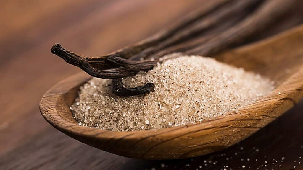 A wooden spoon holds granulated vanilla sugar with two whole vanilla beans resting on top, placed on a wooden surface.