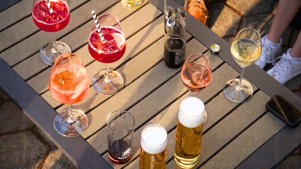 A table with assorted drinks including cocktails, beer, soda, and wine glasses—an inviting spread that reflects classic American BYOB etiquette—pictured outdoors with people’s legs visible nearby.