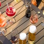 A table with assorted drinks including cocktails, beer, soda, and wine glasses—an inviting spread that reflects classic American BYOB etiquette—pictured outdoors with people’s legs visible nearby.