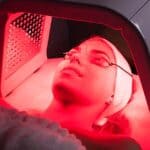 A person wearing protective goggles receives red light therapy on their face, lying under an LED light panel in a clinical setting, providing an insightful look into what is red light therapy and how it is administered.