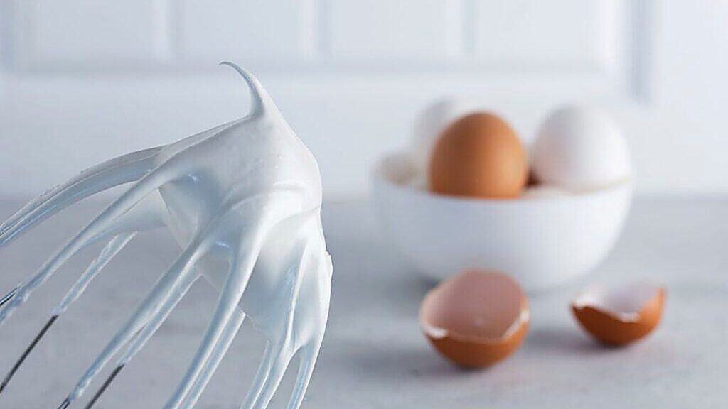 A whisk with stiff egg white peaks in the foreground, and a bowl of whole eggs with eggshells in the background on a white surface.