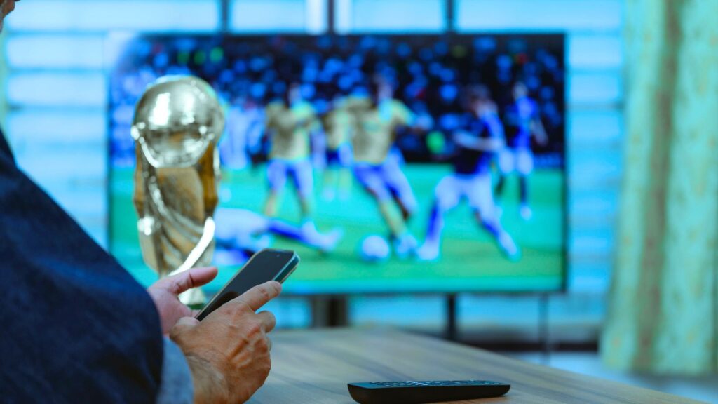 A person holding a smartphone sits by a table with a remote and trophy, enjoying a soccer match on TV in the background via a free World Cup stream.