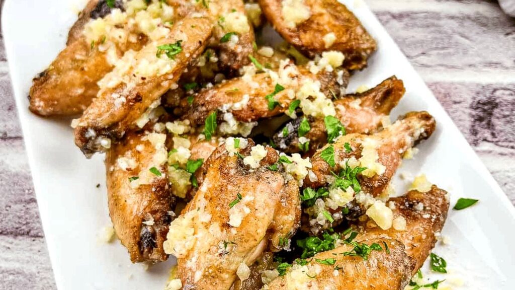A white plate of baked chicken wings topped with minced garlic and chopped parsley.