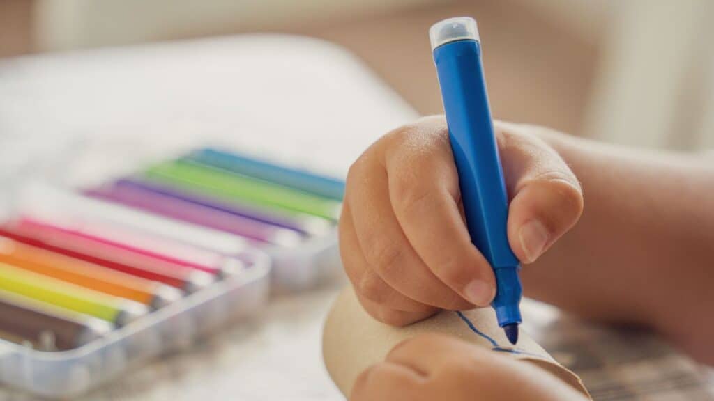 A child&rsquo;s hand draws on a cardboard tube with a blue marker, reminiscent of &rsquo;80s and &rsquo;90s school supplies; a set of colored markers is visible in the background.