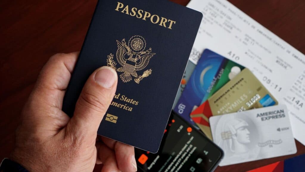 A hand holds a U.S. passport above a table with credit cards, a boarding pass, and a smartphone&mdash;reminding travelers to review travel fees before you fly.