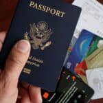 A hand holds a U.S. passport above a table with credit cards, a boarding pass, and a smartphone—reminding travelers to review travel fees before you fly.