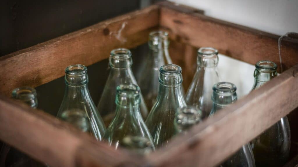 A wooden crate holds twelve empty, clear glass bottles arranged in three rows of four, viewed from above and at an angle, evoking nostalgic milkman delivery memories.