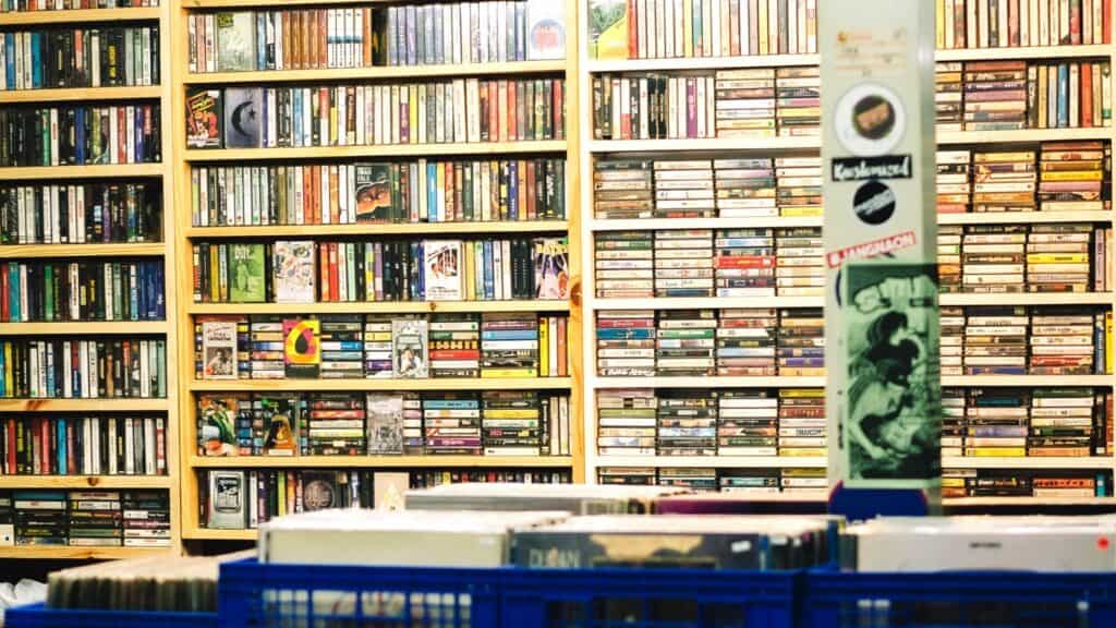 Shelves filled with books and cassette tapes line the wall behind blue crates containing records in a store, evoking the nostalgic charm of video rental chains everyone remembers.