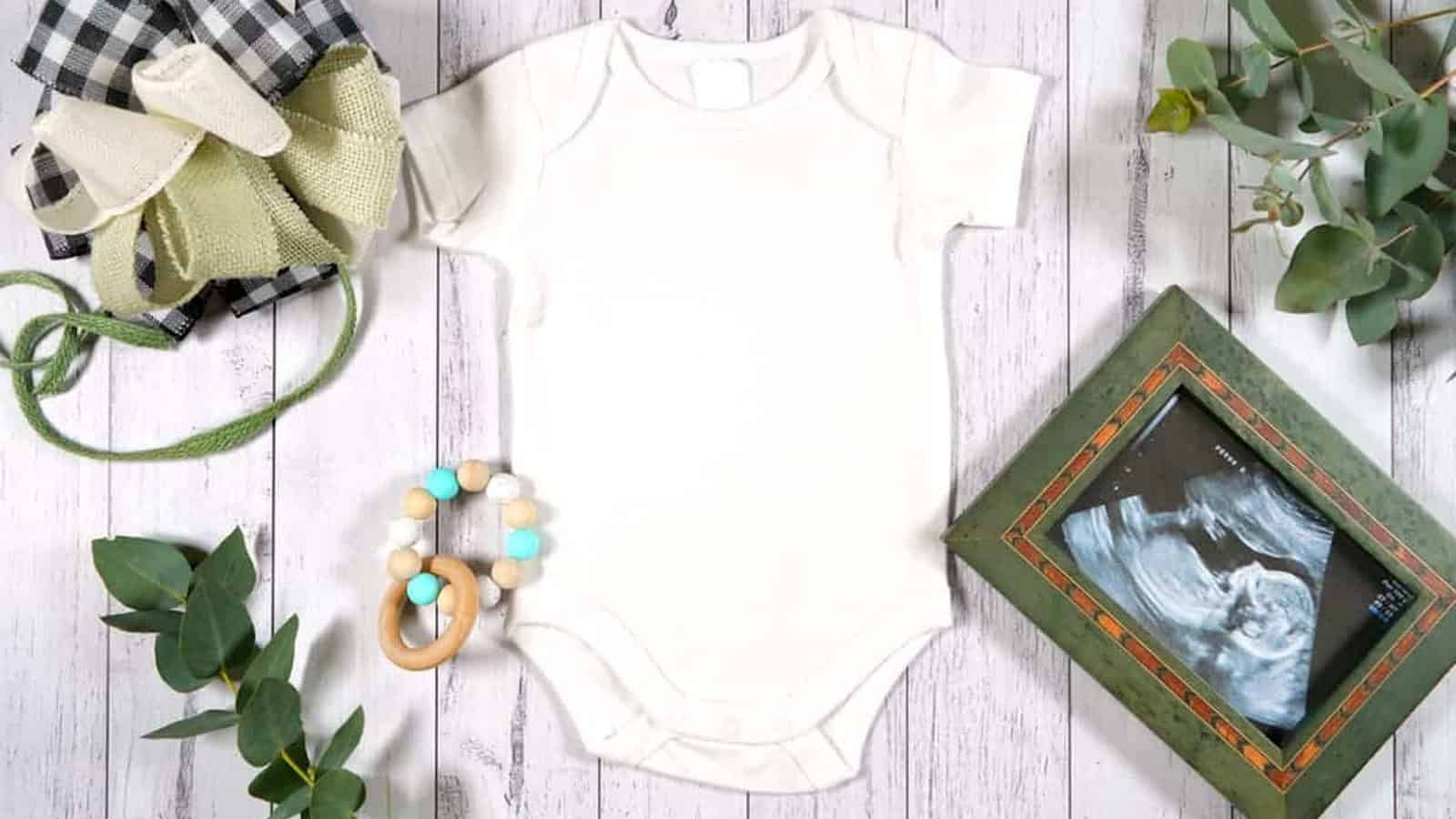 Flat lay of a plain white baby onesie, wooden teether, folded fabric, baby shoes, greenery, and framed ultrasound photo on a white wooden surface—perfect for inspiring creative ideas on how to reveal pregnancy.